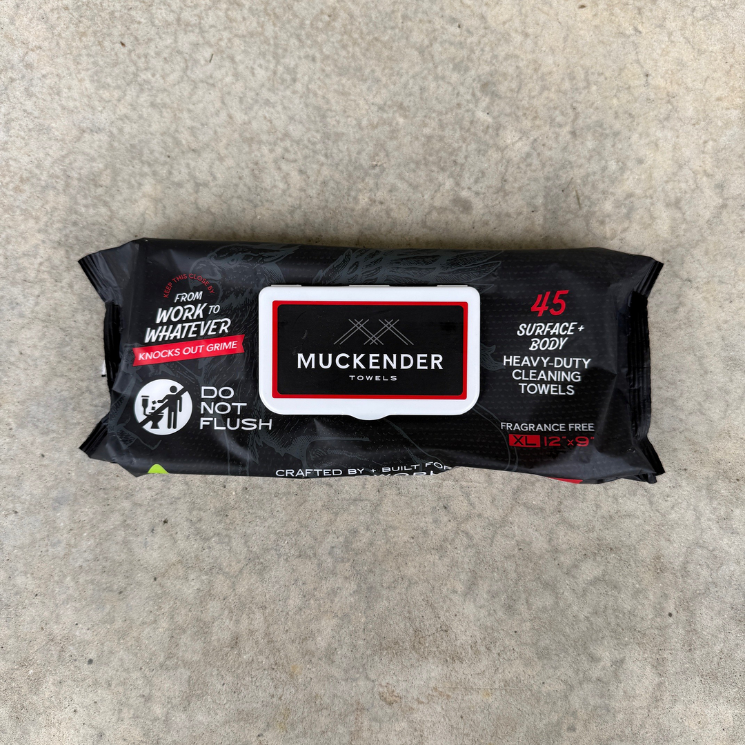 A package of Muckender Towels cleaning towels lies on a textured gray concrete floor. The packaging is black with a white and red central label